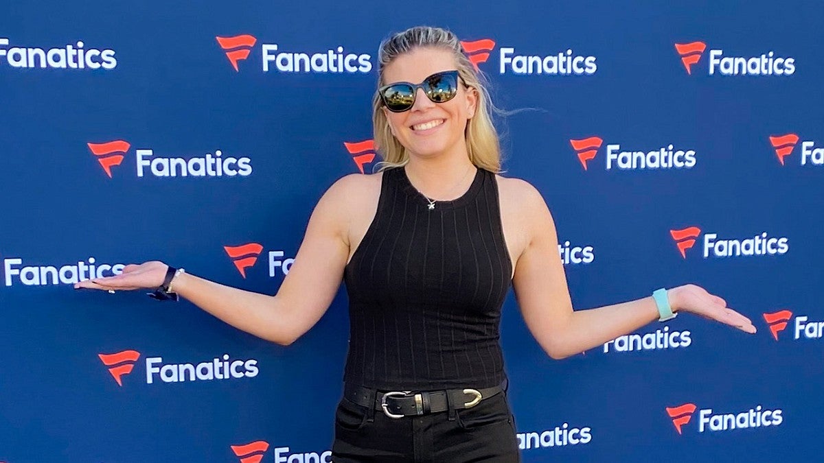 Hannah Oakley wears sunglasses and poses in front of a blue wall covered in the Fanatics logo