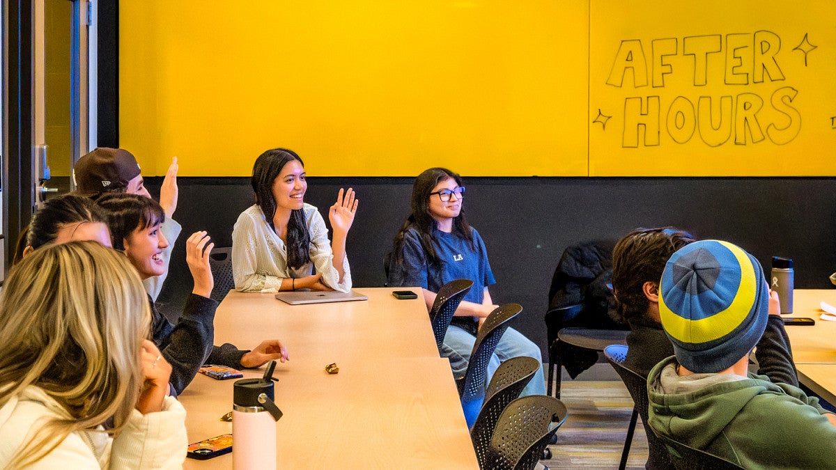 students raise their hands in a classroom in front of a large yellow wall where someone has written "After Hours"