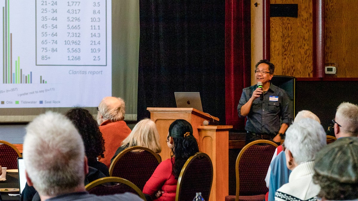 Andrew DeVigal speaks into a microphone in front of a group of seated people looking at a display screen
