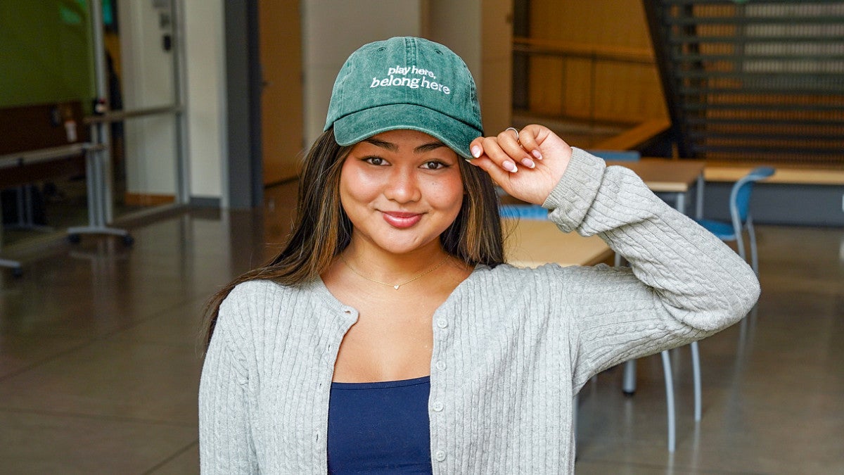 University of Oregon public relations student Sadie Schumaker poses with a baseball cap embroidered with the slogan "Play Here. Belong Here"