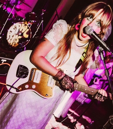 Kayla Krueger plays guitar for her band Grrlpower wearing a white dress and costume makeup