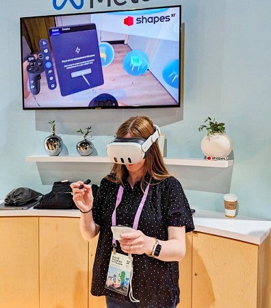 Kathleen Darby wears a VR headset in front of a wall with the Meta logo and a large screen showing an immersive environment