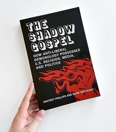 a hand holding a copy of the book The Shadow Gospel by Whitney Phillips and Mark Brockaway