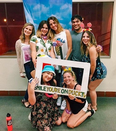 a group of University of Oregon School of Journalism and Communication students, including Hannah Oakley, pose with a cardboard frame that says "Smile, you're on DuckTV"