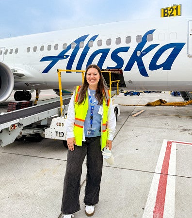 Maya Valverde wears a neon safety vest while posing in front of an Alaska Airlines plane during her summer public relations internship