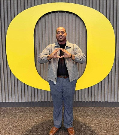 Berto Jolliffe holds his hands in the shape of an O while posing in front of a large yellow University of Oregon logo