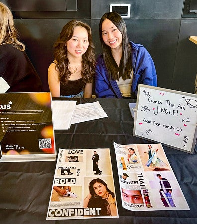 Two students pose while sitting at a table covered in signage for Taking Up Space