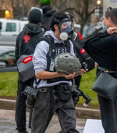 a student photographer wearing a gas mask and carrying a helmet at a protest