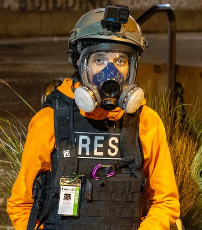 Saj Sundaram poses while wearing a helmet, gas mask, and press vest