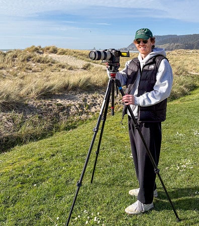 McKain Williams poses with a camera on a tripod, looking over grassy dunes 