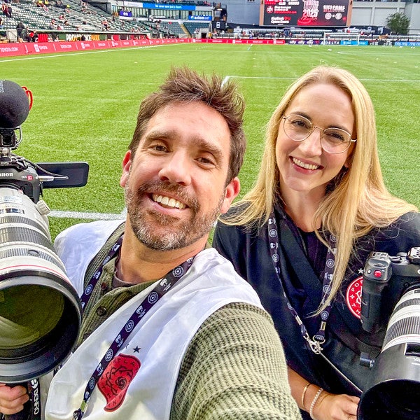 two people take a selfie while shooting video on the sidelines at a professional soccer field