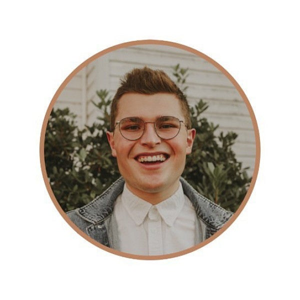 SOJC Alum Maxwell Ely stands, smiling, in front of a white building and foliage. 