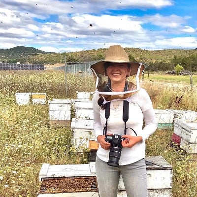 Sierra McClain wears a beekeeper's hat and holds a camera in a field with several beehives