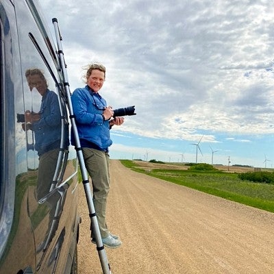 Erika Bolstad stands on a ladder on the side of a van with a camera in her hands