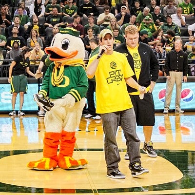 Alex Horwitch speaks into a microphone from the Oregon basketball court while standing next to the Duck mascot