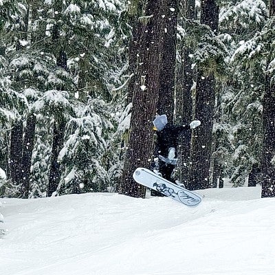 Sydney Seymour performs a jump on a snowboard amid snowy pine trees