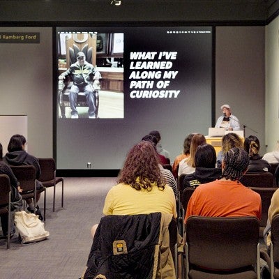 Dave Koranda delivers a lecture to a room full of students in front of a large screen that says "What I've Learned Along My Path of Curiosity"