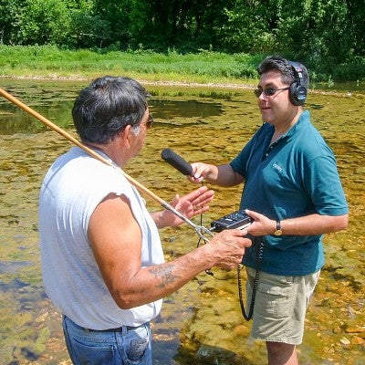 Brian Bull records an audio interview with another person while standing in a shallow murky pond