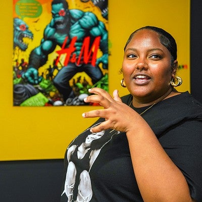 Rachelle Ross stands in front of a yellow wall with a colorful poster