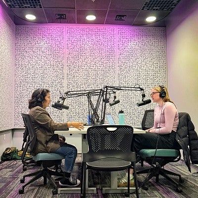 two people sit across from each other wearing headphones and speaking into microphones while recording a podcast
