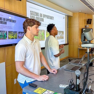 two students present in front of digital screens