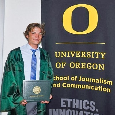 Sam Katzman posing in a graduation gown next to a banner that says University of Oregon School of Journalism and Communication