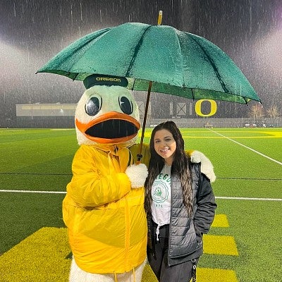 Sarah Ortiz poses with the Oregon Duck under an umbrella while standing on an empty football field in the rain