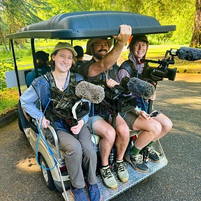 Three people holding video recording equipment on the back of a golf cart