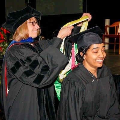 Ammina Kothari wears a black cap and gown and kneels while Leslie Steeves places a yellow stole over her head