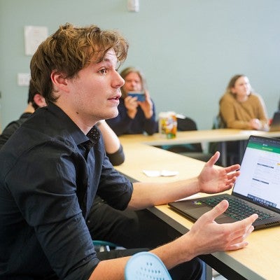 a student in the foreground speaks to someone off camera; other students in the background sit around the table