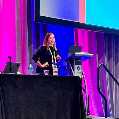 Sara Roth stands at a podium on a stage in front of a backdrop lit in vibrant blues and pinks