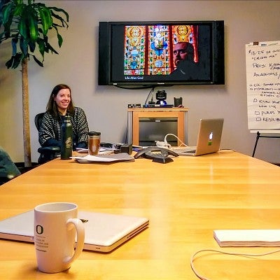 Sara Roth and Wes Pope sit at a large conference table in front of a TV screen displaying a multimedia project 