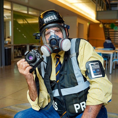 Dan Morrison poses while wearing a press helmet, a gas mask, a press vest, and holding his camera