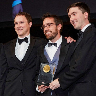 four men wearing tuxedos posing with a glass award trophy
