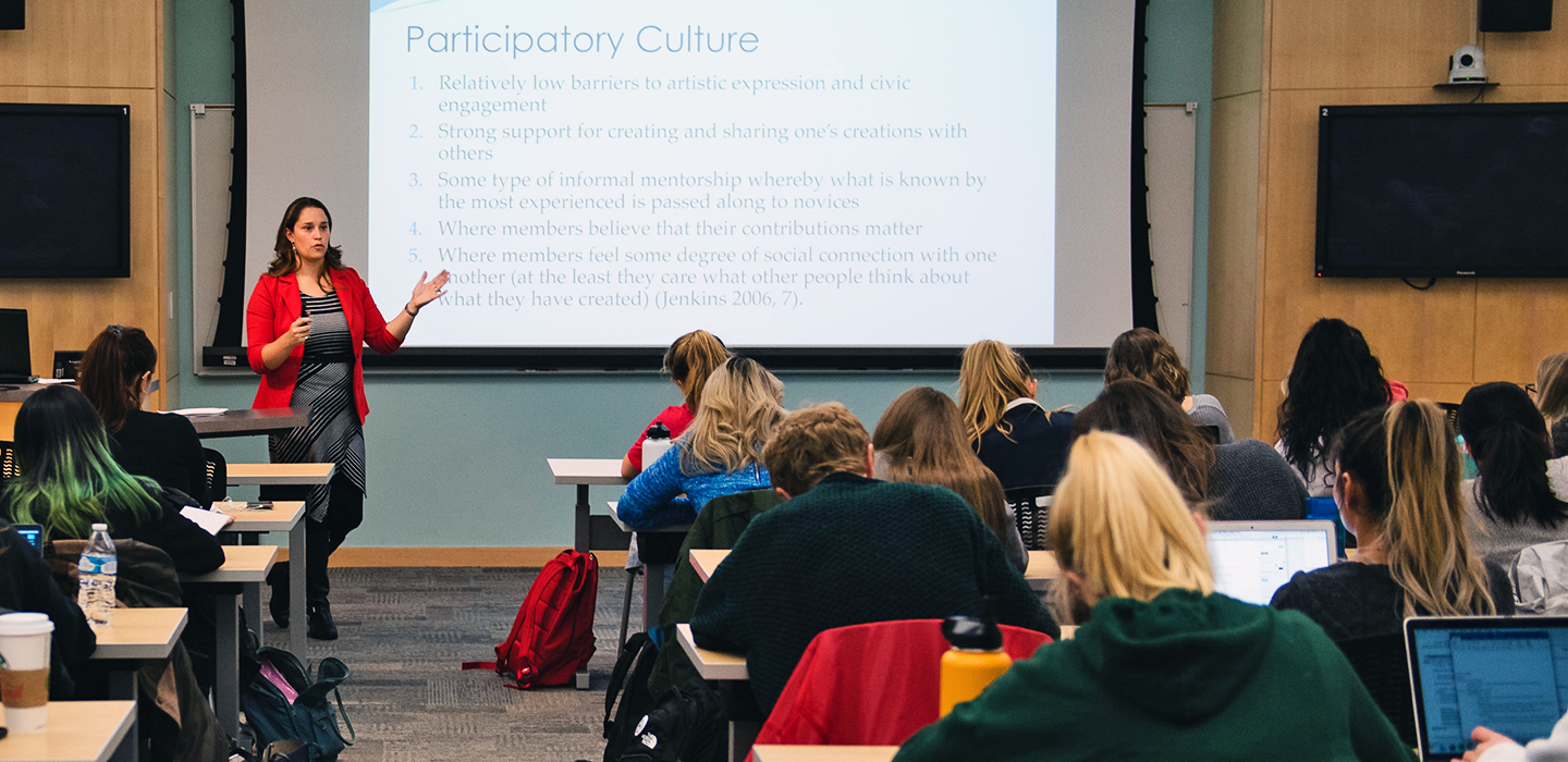 Amanda Cote stands in front of a projector screen that includes the text &quot;participatory culture&quot; while lecturing to a class of students