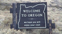 A wooden 'Welcome to Oregon' sign stands in a field, wishing that travelers enjoy their visit.