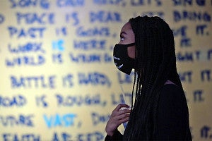 A person wearing a mask stands in profile in front of a wall with handwritten text