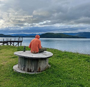 A person wearing an orange jacket sits on a wooden table outside in Cordova, Alaska