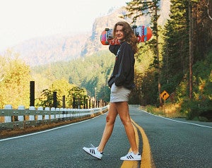 Caitlin Crowley crosses a rural highway carrying a skateboard over her shoulder