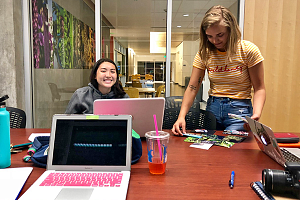 two students work at a table with laptops, a camera, and photos