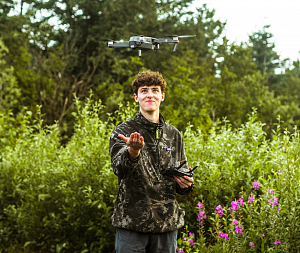 a student with an outstretched hand pilots a drone, which appears to be hovering above their hand. They are standing outside in front of lush green foliage.