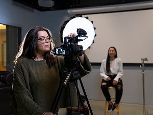 Two students work in the production studio. One student is seated in a stool, while the other student adjusts the settings on video camera. 