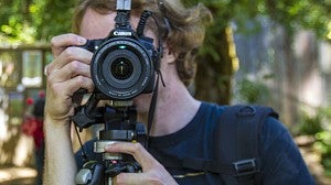 A male student behind a digital camera on a tripod looking through the viewfinder