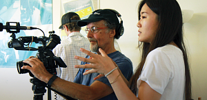 A person wearing a black baseball cap operates a video camera on a tripod while a person next to him in a white shirt gestures