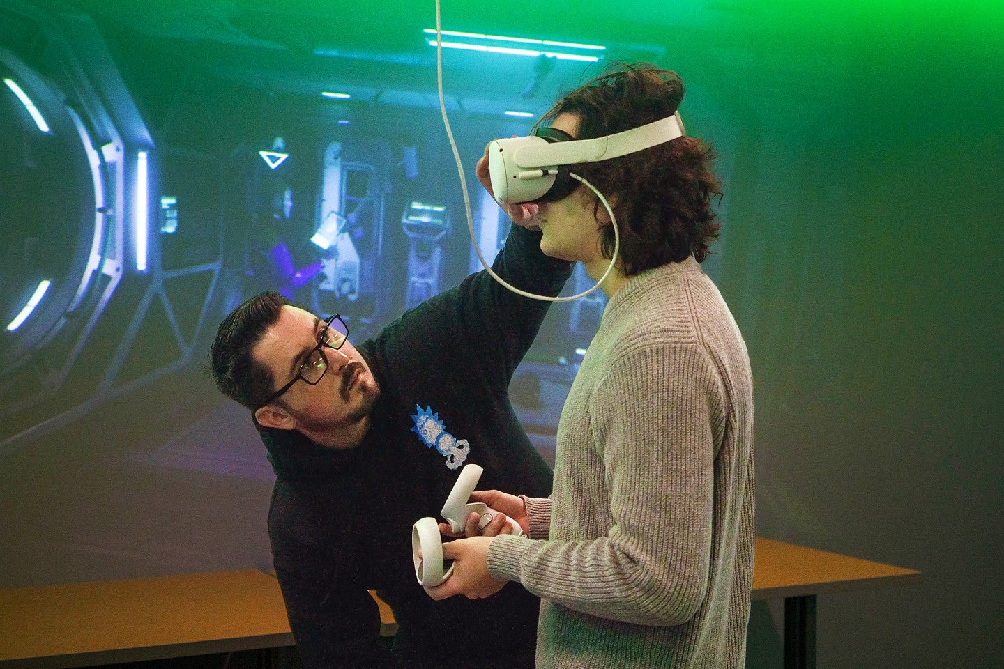 two people work with VR equipment in front of a screen displaying the VR content