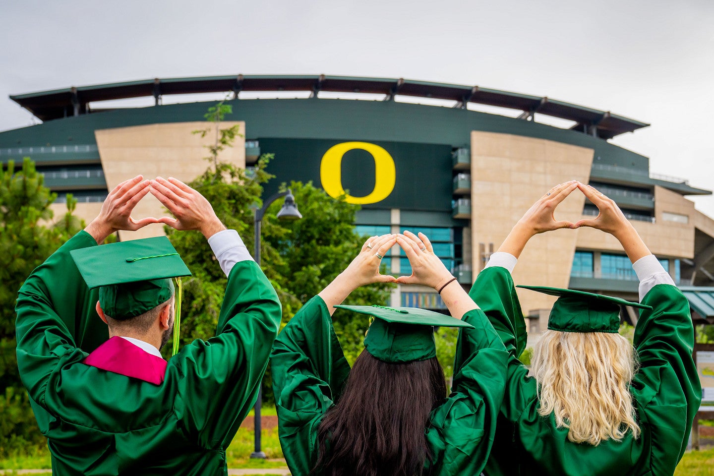 Three students wearing green graduation caps and gowns throw the O in front of Autzen Stadium