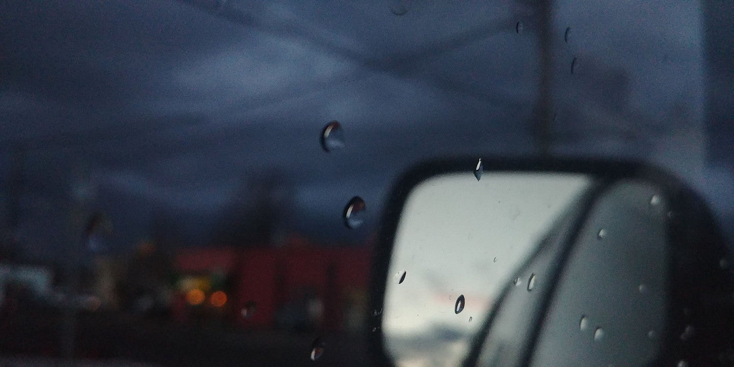 moody photo of raindrops on a car window in focus, with a blurry car mirror and landscape in the background