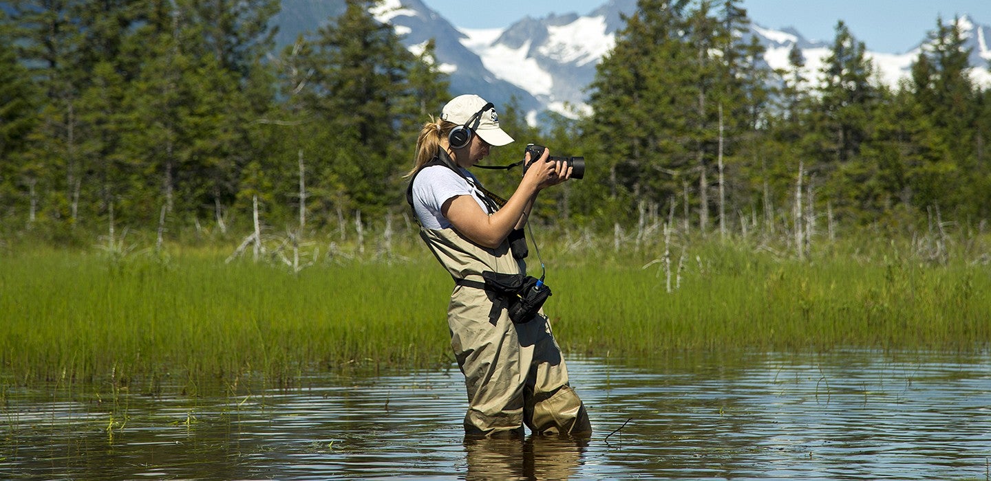 A female student in waders standing in knee deep water fiming on a digital camera