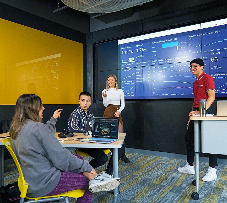 Social Media Lab in the SOJC Experience Hub group of four UO students have a discussion in a classroom setting, with a large screen showing charts and graphs behind them