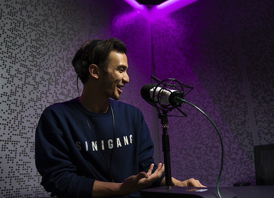One student is seated in the podcast studio in front of a microphone. They are illuminated by a purple light. 